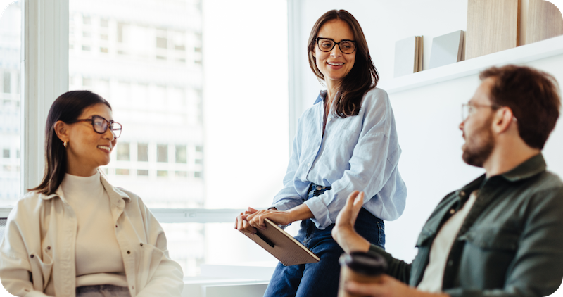 A woman with a clipboard and glasses speaks to 2 colleagues in a bright conference room